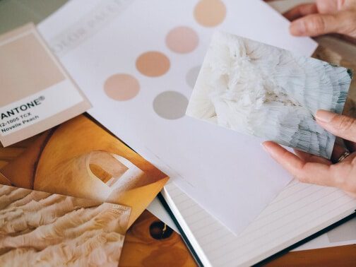 A person holds a marbled white sample card over a desk with color swatches, samples, and a Pantone color book, suggesting design or interior planning work.