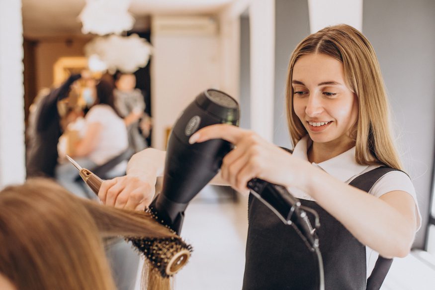 Hair stylist blow‑drying a client’s hair with a round brush in a bright, modern salon.