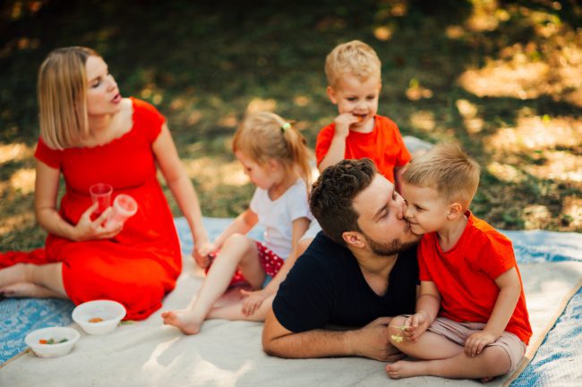 Family picnic in a sunny park with a mother in a red dress, father kissing their son, and siblings snacking on a blanket, evoking warm, reliable outdoor moments