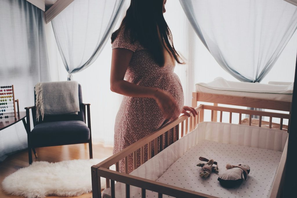 Expectant mother standing by a wooden crib in a sunlit nursery