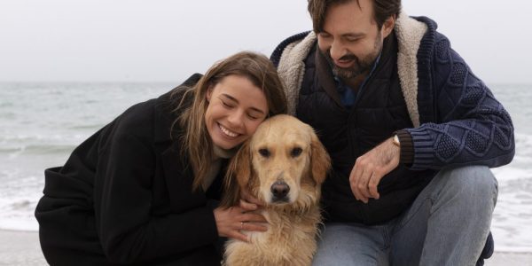 Couple kneeling on a windswept beach beside their golden retriever at sunset, illustrating the emotional bond fueled by reliable pet nutrition and travel-friendly feeding gear