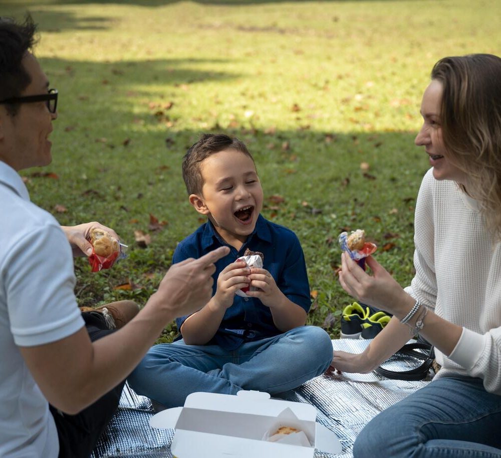 Parents and young son laughing over snacks on a picnic blanket in the park, symbolizing the family time unlocked by hassle-free auto-refill pet subscriptions