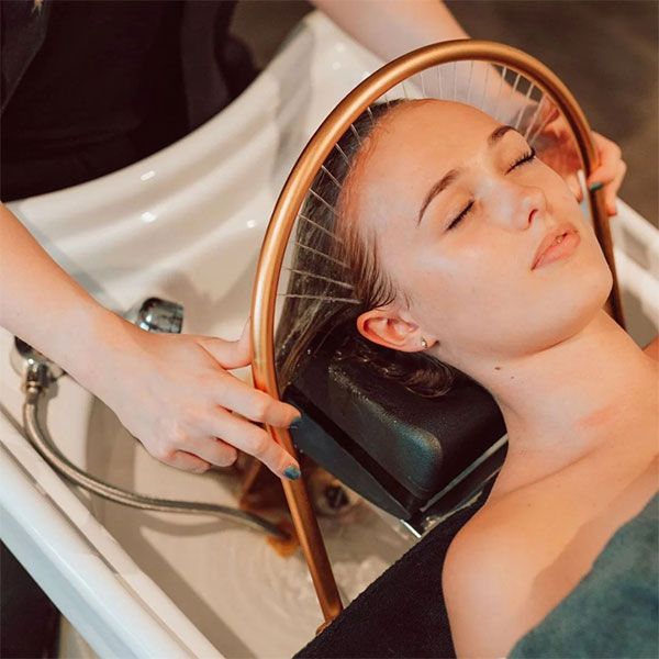 Stylist brushing and blow‑drying a client’s hair using a round brush and hairdryer in a bustling salon.