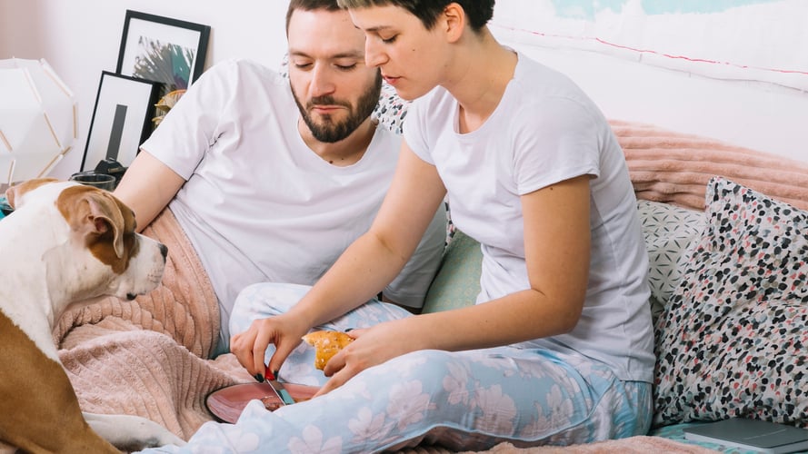 Couple feeding their dog by hand on a bed, highlighting manual workarounds when automatic food subscriptions fail