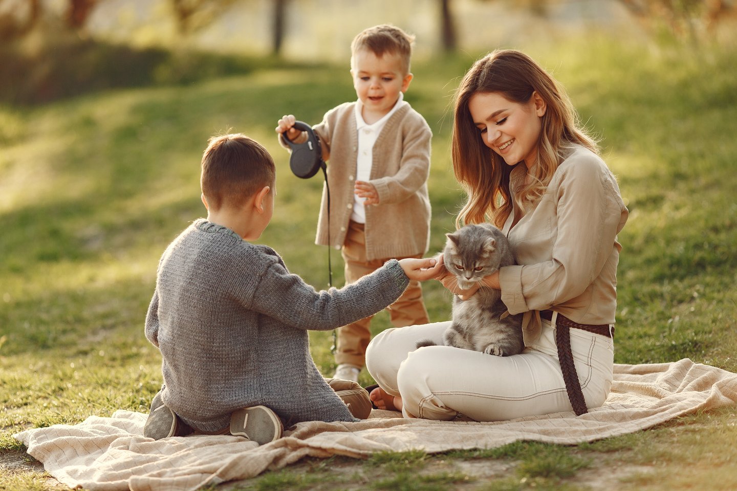 Young mother seated on a picnic blanket outdoors holding a gray cat as two boys, one with a leash reach in to pet it in soft afternoon light