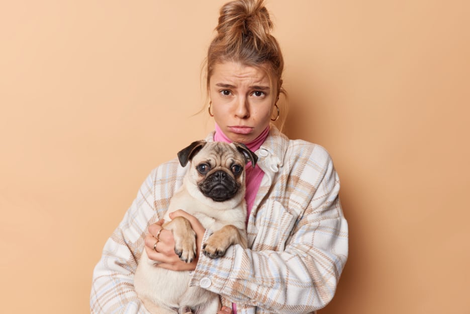 Worried woman holding a small pug against a solid background, conveying frustration with clunky pet subscription tools
